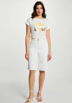 Donna che indossa una maglietta bianca con grafica e la scritta "The Sun Comes Up", pantaloncini bianchi fino al ginocchio e sandali bianchi con tacco a blocco e cinturini, in piedi.