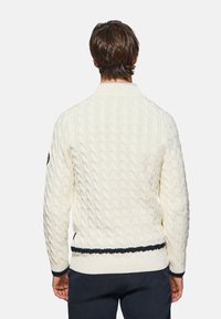 Suéter blanco de punto trenzado con cuello alto, dobladillo a rayas en azul marino y puños acanalados. Incluye un detalle de parche en la manga izquierda.