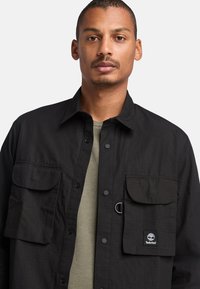 Giacca camicia nera con bottoni a pressione, dotata di due tasche frontali, realizzata in tessuto texture. Presenta un logo Timberland e un dettaglio in metallo circolare.