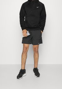 Svart Nike-hoodie med framficka och dragkedjedetalj, tillsammans med mörka shorts och svarta sneakers. Vattenflaska hålls i vänster hand.