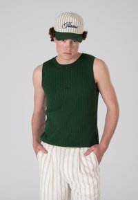 Green ribbed sleeveless top with a crew neckline, paired with beige and cream striped trousers. The model wears a cream cap with green accents.