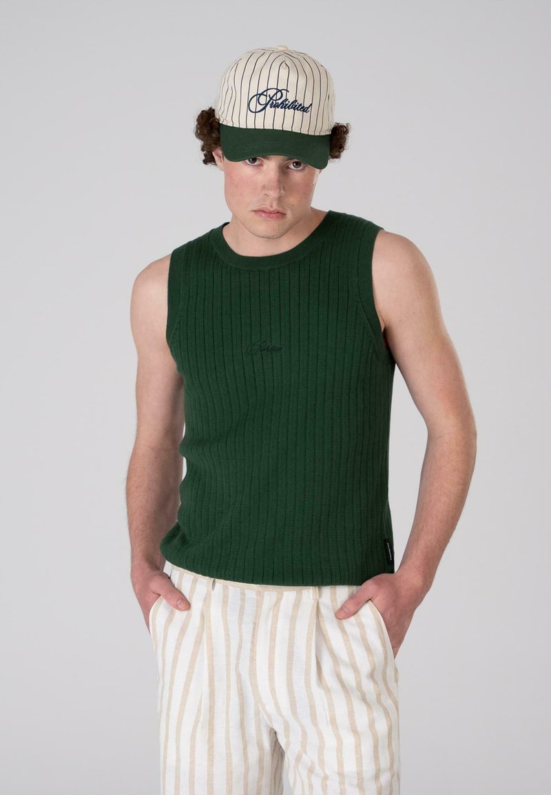 Haut sans manches vert à côtes avec un col rond, accompagné de pantalons rayés beiges et crème. Le modèle porte une casquette crème avec des accents verts.