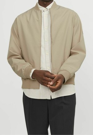 Homme portant une veste bomber beige sur une chemise beige et blanche à rayures, avec un pantalon noir, les mains jointes légèrement devant.