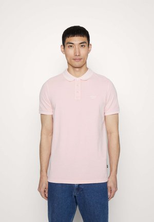 Polo de algodón en color rosa claro, con tapeta de dos botones, mangas cortas y un pequeño logo en el pecho. Combinado con unos vaqueros azules.