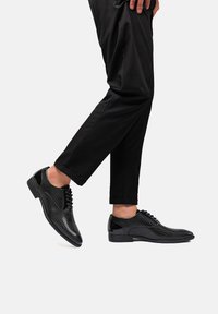 Scarpe eleganti nere con superficie testurizzata, design con lacci e dettagli lucidi. Abbinate a pantaloni neri a sigaretta, che evidenziano un profilo snello.