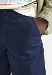 Los pantalones de algodón azul marino presentan un frente plano y bolsillos laterales, combinados con una chaqueta verde claro y un suéter de punto a rayas.