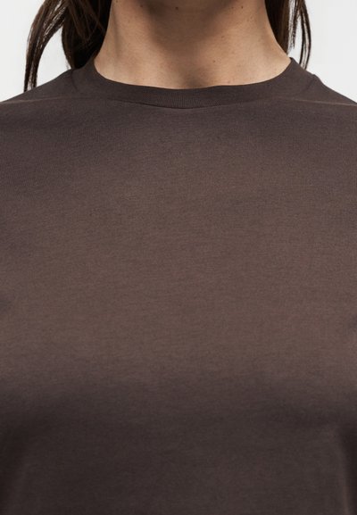 Κοντινό πλάνο ενός ατόμου που φοράει απλό καφέ μπλουζάκι με στρογγυλή λαιμόκοψη και μακριά σκούρα μαλλιά ορατά πίσω από τους ώμους.