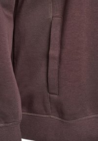Sudadera marrón con una textura suave, que cuenta con un bolsillo lateral y puños acanalados. Detalle de costura visible a lo largo del dobladillo y las costuras.