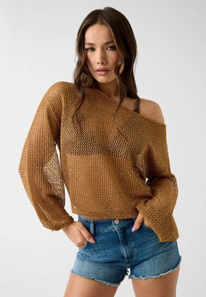 Jonge vrouw draagt een losse bruine mesh trui die van één schouder is gevallen en blauwe spijkershorts, poserend met handen in de zakken tegen een effen achtergrond.