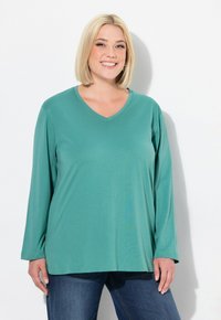 Türkisfarbenes Langarm-V-Ausschnitt-Shirt aus weichem Stoff, mit lockerer Passform und schlichten Design, kombiniert mit blauen Jeans.