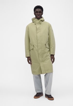 Mand iført en lysegrøn lang hættejakke, lyseblå jeans og mørke sneakers, står mod en hvid baggrund.