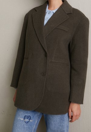 Personne portant un blazer marron foncé boutonné par-dessus une chemise bleu clair à col et un jean bleu déchiré avec un patch graphique.