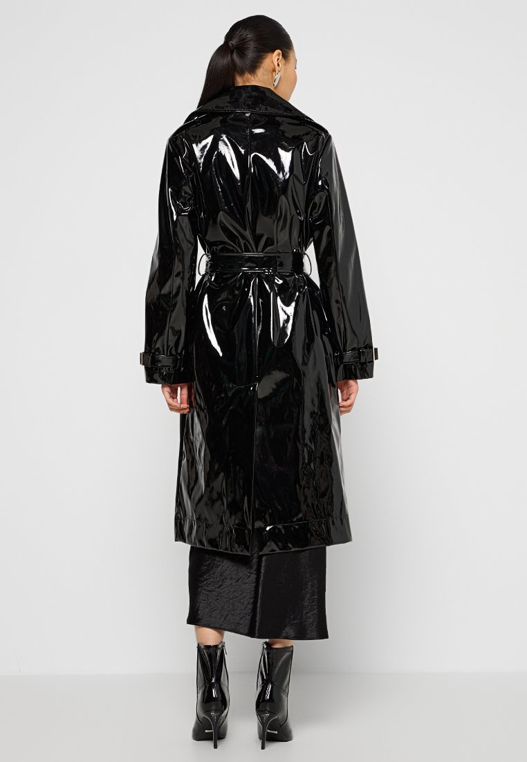 Schwarzer Lackleder-Trenchcoat mit tailliertem Schnitt, langen Ärmeln und glänzender Oberfläche, kombiniert mit einem schwarzen Rock und Stiefeln mit Absatz.