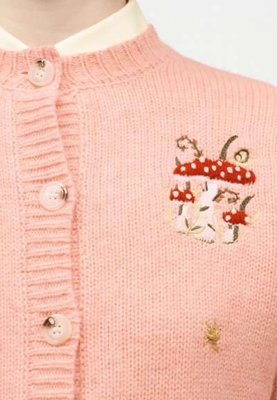 Ροζ πλεκτή ζακέτα με μπεζ κουμπιά, διακοσμημένη με κεντητά κόκκινα και λευκά μανιτάρια με πράσινα φύλλα στο στήθος.