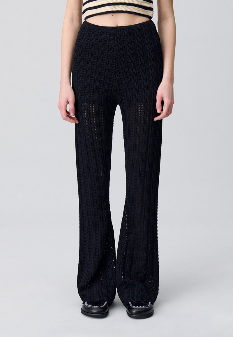 Pantalon large en tricot noir avec texture côtelée et tissage ouvert. Taille élastique, longueur au sol et chevilles évasées. Design simple, sans motif.