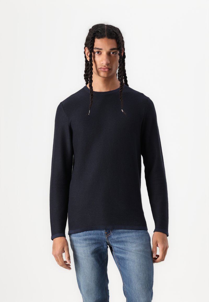 Junge Person mit langen, geflochtenen Haaren, die ein dunkelblaues Langarmshirt und eine hellblaue Jeans trägt, steht vor einem weißen Hintergrund.