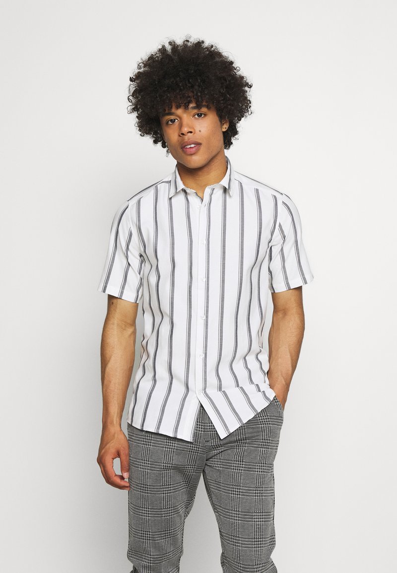 Short-sleeve button-up shirt in white with vertical black stripes, featuring a pointed collar and a straight hem, paired with checkered trousers.