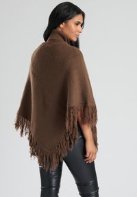 Brun stickad poncho med hög krage, med fransade kanter och en strukturerad mönster. Bärs över svarta åtsittande byxor.