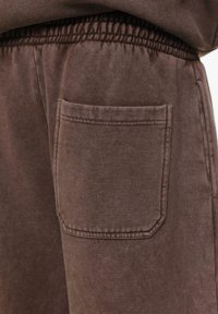 Bruine sweatpants hebben een gestructureerde stof, een elastische tailleband en een enkele opgestikte zak aan de zijkant. De stiksels zijn zichtbaar rond de zak.