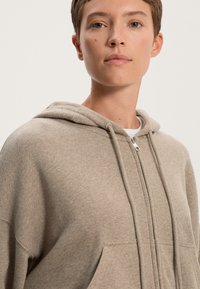 Felpa beige con zip realizzata in tessuto morbido e testurizzato. Presenta un cappuccio, lacci e tasche frontali. Design minimalista senza pattern.