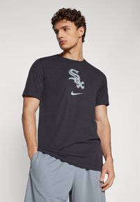 T-shirt de algodão preto com um gráfico "Sox" em cinza e branco, com um logótipo da Nike abaixo. Modelo a usar calções cinza claro, com as mãos nos bolsos.
