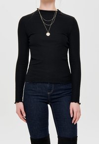 Haut noir côtelé à manches longues avec un col montant, assorti à un jean bleu foncé. Comprend des colliers dorés superposés et des bords de manches effilochés.