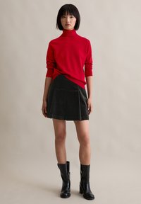 Roter Rollkragenpullover, schwarzer plissierter Cordrock und kniehohe schwarze Stiefel mit Absatz, vor einem neutralen Hintergrund stehend.