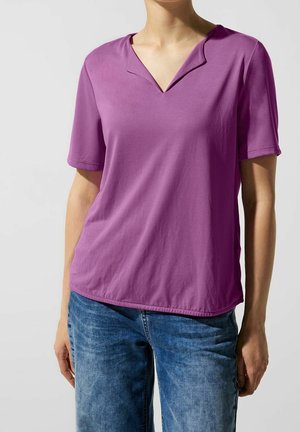 Person iført en lilla kortærmet top med V-udskæring og blå denimjeans, stående mod en ensfarvet lys baggrund.