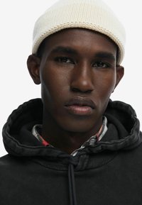 Primo piano di una persona che indossa un berretto di lana bianco e una felpa nera con cordino. È visibile un colletto che presenta una camicia a scacchi sotto.