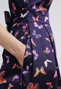 Kvinnas hand i en marinblå klänning ficka med tryck av rosa, lila och orange fjärilar samt en bred midjeband med en rosett på sidan.