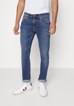 Hombre con vaqueros ajustados azules remangados en los tobillos, zapatillas blancas con detalles rojos y una camiseta blanca, de pie frente a un fondo liso.