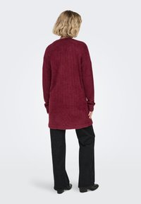 Bordeauxfarvet blød lang cardigan med tekstureret stof, sænkede skuldre og sidelommer, kombineret med sorte flared bukser og ankelstøvler.