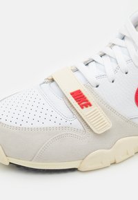 Sneaker bianca con tomaia in suede e pelle, logo "NIKE" rosso, punta della scarpa perforata, cinturino elastico. Suola in gomma spessa con trazione nera.