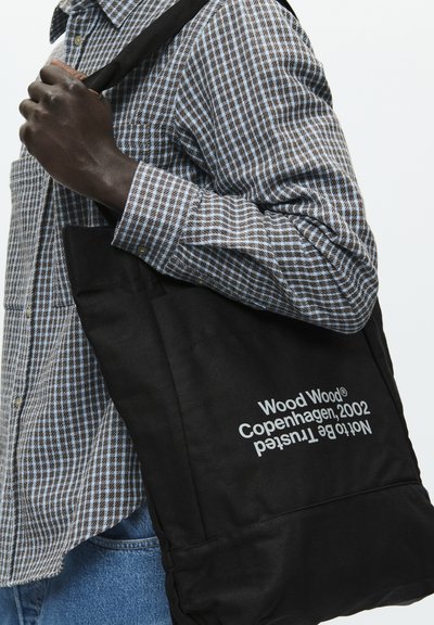 Fekete vászon táska téglalap alakú, fehér felirattal, masszív vállpánttal, egy kockás ing mellett hordva.