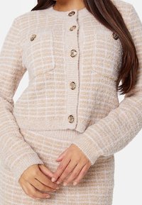 Cardigan beige clair avec un motif texturé à carreaux. Il comporte des boutons dorés, deux poches avant et des poignets et un ourlet côtelés.