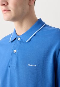 Modrá polo košile vyrobená z texturované tkaniny, s klasickým límečkem, bílými akcentními pruhy a malým vyšitým logem "GANT" na hrudi.