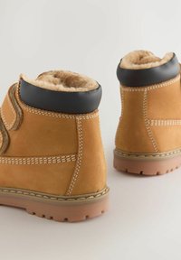 Tan-farbene Wildleder-Stiefeletten mit einem gepolsterten Kragen in Schwarz, weißen Nähten und Gummisohlen. Mit weichem Fell gefüttert für zusätzliche Isolierung.