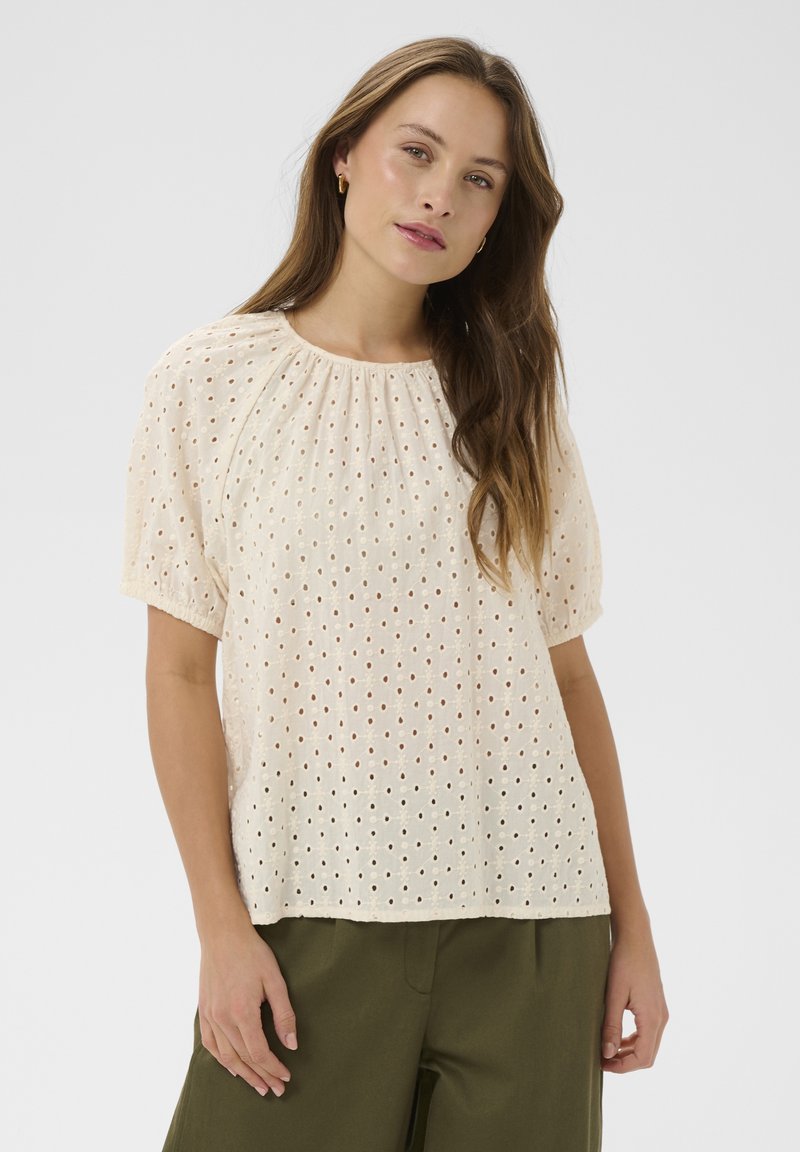 Jeune femme aux longs cheveux bruns portant un chemisier crème à manches courtes ajouré et un pantalon vert olive, debout devant un fond clair.