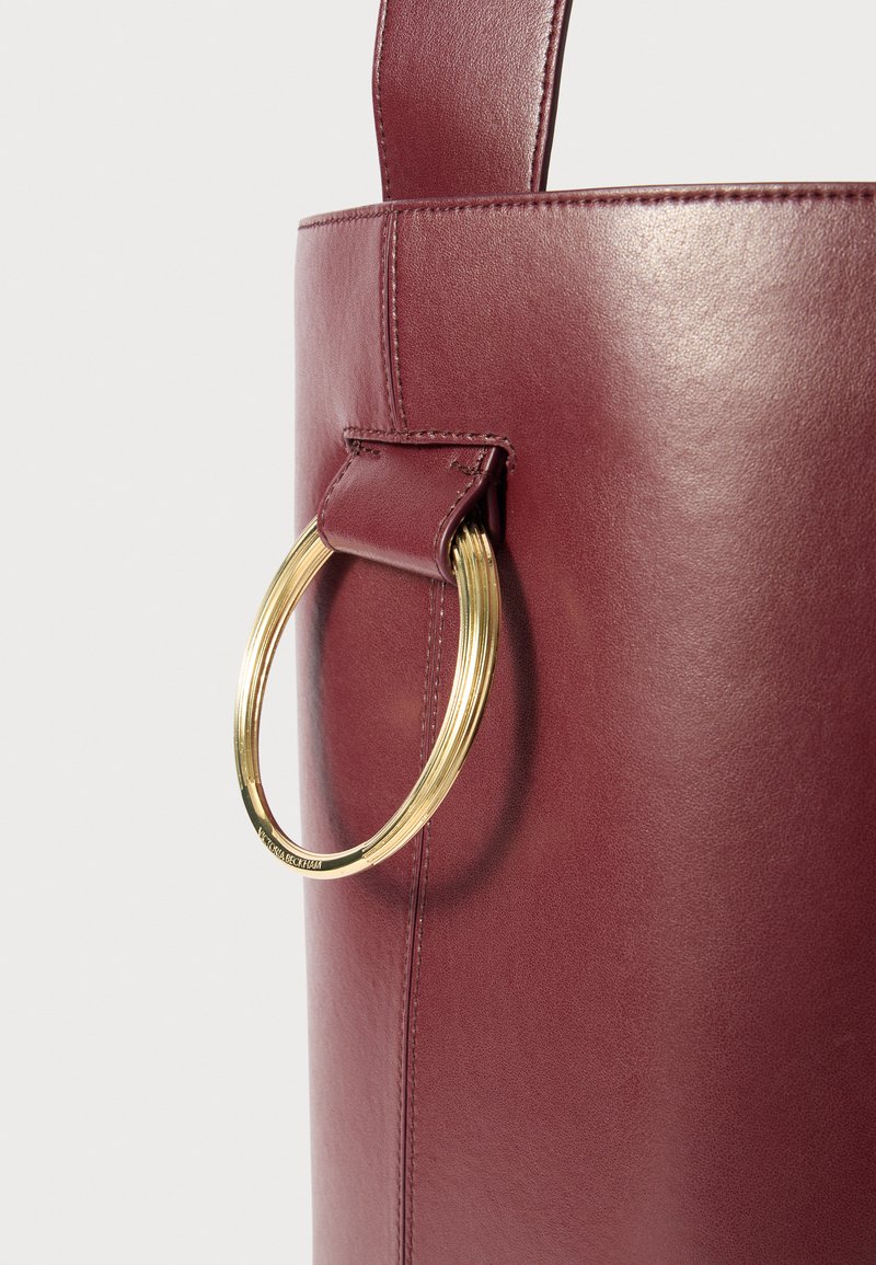 Sac en cuir bordeaux avec une texture lisse, doté d'un accent en métal doré et d'un attachement de poignée cousu. Forme cylindrique.