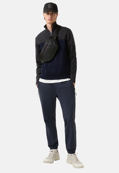 Navy- und grauer Zip-Kragen-Pullover mit gerippter Textur, navy Jogginghosen, schwarze Umhängetasche und beigefarbene Sneakers mit dicker Sohle.