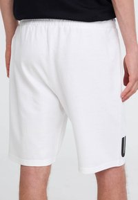 Shorts bianchi in cotone con vita elastica, tasche laterali e un accento grafico nero sulla gamba sinistra. Texture liscia, lunghezza al ginocchio.