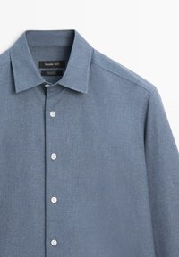 Modrá bavlněná košile se špičatým límečkem, bílými knoflíky a texturovanou látkou. Štítek obsahuje název značky a informace o složení látky.