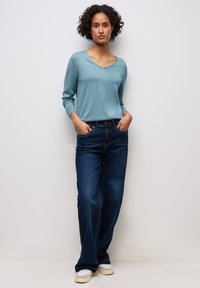 Ljusblå v-ringad tröja med ribbat avslut, ihop med mörkblå vida jeans. Modellen står med händerna i fickorna och har på sig ljusa skor.