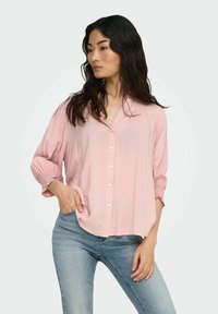Lys pink knaplukket skjorte med V-udskæring, trekvartærmer og sammentrukne manchetter. Båret med blå jeans, der har en afslappet pasform.