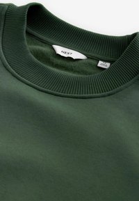 Sudadera de color verde oscuro con cuello redondo acanalado. Textura suave con una etiqueta visible que dice "NEXT" y "MADE IN" cerca del cuello.