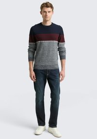 Gestrickter Pullover mit marineblauen, burgunderfarbenen und grauen Farbblöcken, strukturiertem Stoff, Rundhalsausschnitt und langen Ärmeln, kombiniert mit dunklen Jeans und weißen Turnschuhen.
