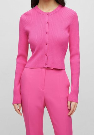Femme portant un cardigan ajusté boutonné rose vif côtelé et un pantalon taille haute assorti, debout devant un fond uni.