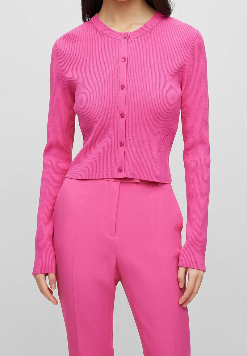 Femme portant un cardigan ajusté boutonné rose vif côtelé et un pantalon taille haute assorti, debout devant un fond uni.