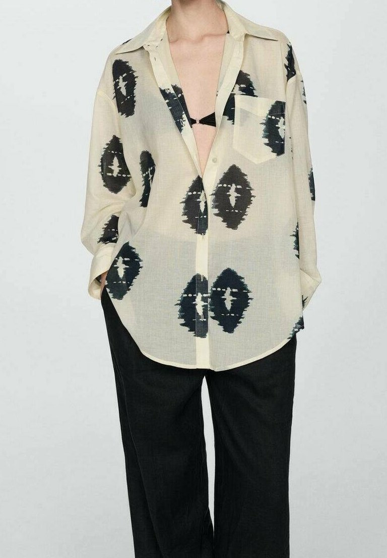 Personne portant une chemise beige oversize avec de grands motifs abstraits noirs sur un pantalon noir, chemise partiellement ouverte révélant un haut noir en dessous.