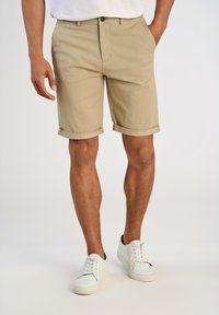 Shorts en coton beige avec un ourlet roulé. Poches avant et fermeture par un seul bouton. Portés avec des baskets blanches, se tenant sur un fond neutre.